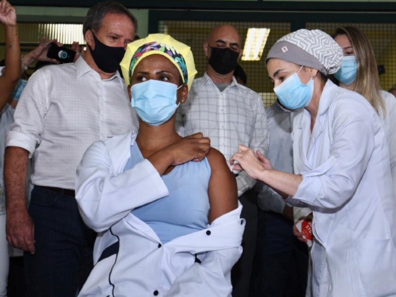 Em primeiro plano, a técnica de enfermagem recebe a vacina de uma enfermeira no braço esquerdo. Ela é negra e usa uma toca amarela com faixa florida no cabelo e está de máscara